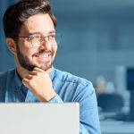 Ein fröhlicher junger Mann mit Bart und Brille, der nachdenklich vor einem Laptop in einem modernen Büro sitzt.
