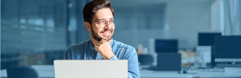 Ein fröhlicher junger Mann mit Bart und Brille, der nachdenklich vor einem Laptop in einem modernen Büro sitzt.