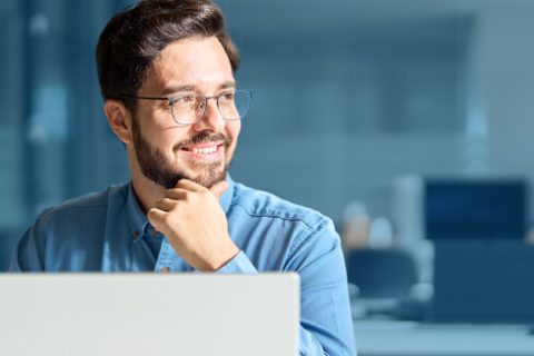Ein fröhlicher junger Mann mit Bart und Brille, der nachdenklich vor einem Laptop in einem modernen Büro sitzt.