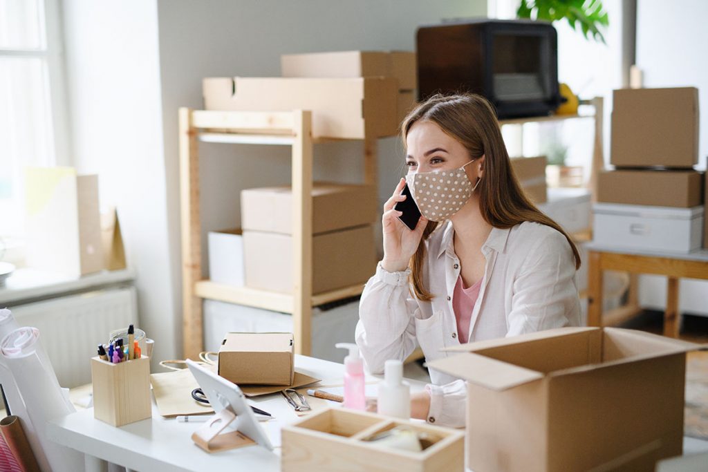 Frau mit Mundschutz telefoniert in ihrem Lager des Onlineshops