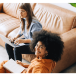 Zwei Frauen arbeiten gemeinsam in einem hellen Raum, eine sitzt auf dem Boden und schreibt in ein Notizbuch, während die andere auf einem Laptop tippt