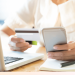 Person, die Online-Zahlung für Domainregistrierung durchführt, hält Kreditkarte in der einen Hand und Smartphone in der anderen. Im Hintergrund ein geöffnetes Laptop und ein Notizbuch mit Stift auf einem Holztisch, symbolisch für die verschiedenen Zahlungsmodelle und Plattformen beim Kauf oder der Miete einer Domain