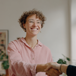 Zwei Geschäftsfrauen schütteln sich die Hände bei einem Geschäftstreffen. Beide lächeln, was auf eine erfolgreiche Verhandlung im B2B-Bereich hinweist.