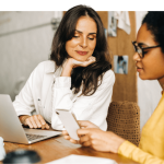 Zwei Frauen bei einer Besprechung, eine nutzt einen Laptop, die andere ein Smartphone.