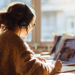 Person in gelbem Pullover hält Kopfhörer vor einem Laptop, mit Text “Das Webinar zur E-Rechnung” im Vordergrund.