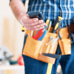 Nahaufnahme eines Bauarbeiters mit Werkzeuggürtel, der auf einer Baustelle sein Smartphone überprüft. Der Bauarbeiter hält einen gelben Schutzhelm und könnte gerade eine Abschlagsrechnung am Handy einsehen oder verwalten.