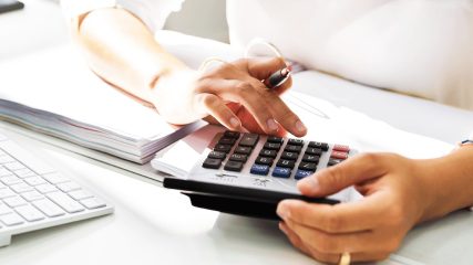 Nahaufnahme einer Person, die mit einem Taschenrechner arbeitet, während sie Zahlen berechnet und Notizen macht.