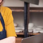 Junge Frau mit Brille und gelbem Shirt, die in einer Werkstatt am Laptop arbeitet und Notizen macht, umgeben von Arbeitsmaterialien