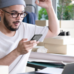 Ein junger Unternehmer sitzt inmitten von Kartons und Verpackungsmaterialien vor einem Laptop und freut sich über eine Bestellung auf seinem Smartphone. Er betreibt ein Dropshipping-Geschäft und verwaltet seine Bestellungen online. Im Hintergrund sind Kleidung und Schuhe für den Versand vorbereitet.