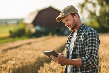 Landwirt steht im Obstgarten und arbeitet auf einem Laptop, neben einer Kiste voller frisch geernteter Äpfel.