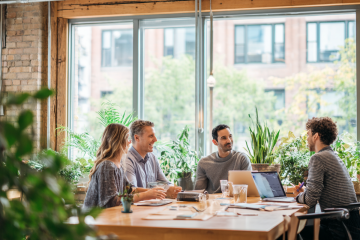 Team diskutiert an einem Laptop über elektronische Rechnungsstellung und EN 16931 in einem modernen Büro. Eine Person macht sich Notizen, während eine andere auf den Bildschirm zeigt, mit Kollegen im Hintergrund.